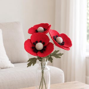 May include: A bouquet of three handcrafted felt poppy flowers with vibrant red petals, black centers, and light beige spherical centers. The flowers feature textured green felt leaves on thin stems and are arranged in a clear glass bottle vase.