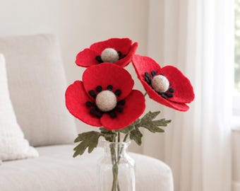 May include: A bouquet of three handcrafted felt poppy flowers with vibrant red petals, black centers, and light beige spherical centers. The flowers feature textured green felt leaves on thin stems and are arranged in a clear glass bottle vase.
