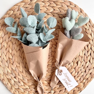 May include: Two bouquets wrapped in brown paper. One bouquet features light green felt eucalyptus leaves. The second bouquet contains light green and grey felt hearts on stems, tied with a white gift tag with the handwritten text Joyeux No&euml;l!