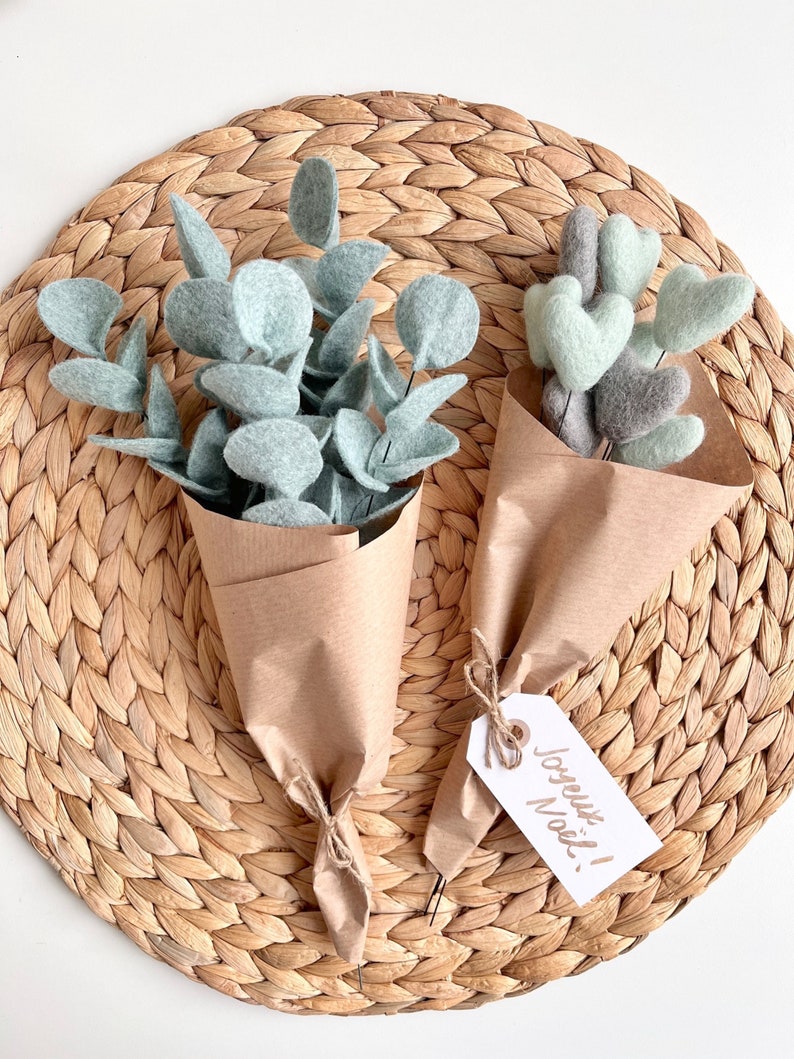 May include: Two bouquets wrapped in brown paper. One bouquet features light green felt eucalyptus leaves. The second bouquet contains light green and grey felt hearts on stems, tied with a white gift tag with the handwritten text Joyeux No&euml;l!