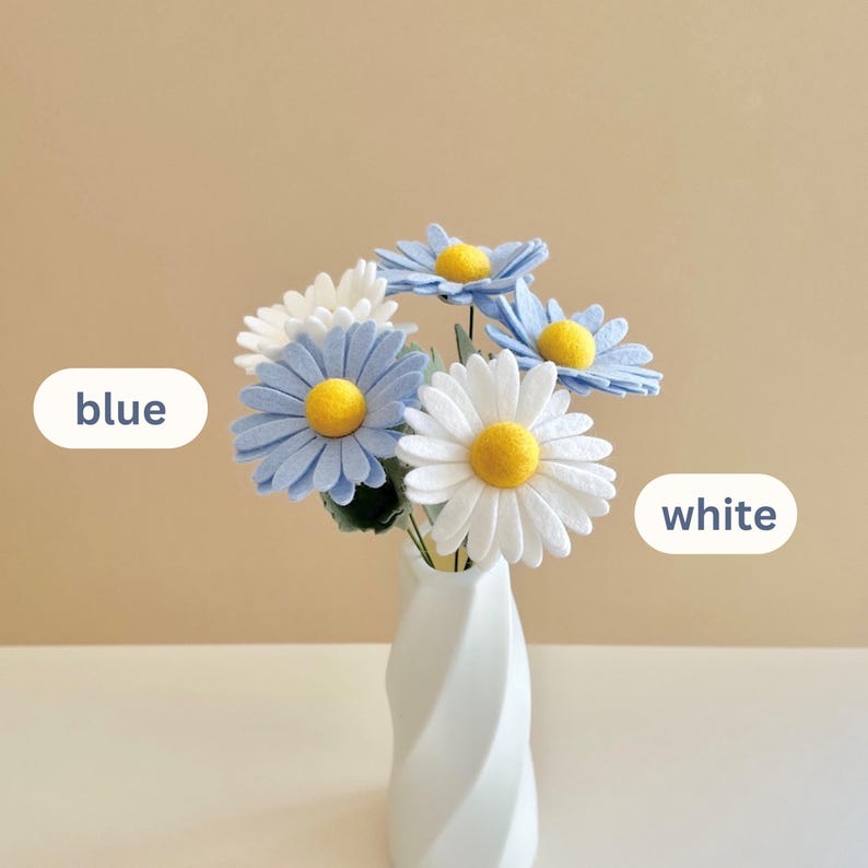May include: A bouquet of felt flowers in a white twisted vase. The flowers feature blue and white petals with bright yellow centers. The image includes text labels pointing to the flowers, with the word blue on the left and white on the right.