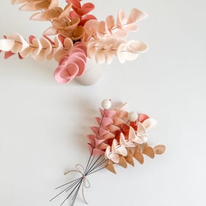 Pode incluir: Um conjunto de ramos de eucalipto em feltro em tons de creme, p&ecirc;ssego e rosa seco. Um grupo est&aacute; disposto num vaso de cer&acirc;mica claro, enquanto o outro &eacute; um ramo solto atado com fio fino, com pequenas bolas de feltro branco em hastes de arame.