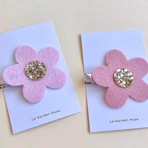 May include: Two hair clips featuring light pink felt flowers with gold glitter centers. Each flower is attached to a silver metal clip mounted on a white display card printed with the text Le Garden Muse at the bottom.