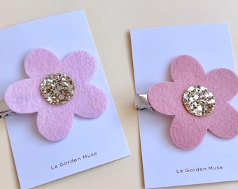 May include: Two hair clips featuring light pink felt flowers with gold glitter centers. Each flower is attached to a silver metal clip mounted on a white display card printed with the text Le Garden Muse at the bottom.