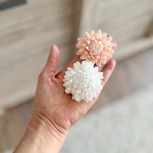 Op de afbeelding: Twee handgemaakte viltbloemen, &eacute;&eacute;n in een zachte perzikkleur en &eacute;&eacute;n in wit, elk met meerdere gelaagde bloemblaadjes in een cirkelvormig patroon die lijken op een dahlia of chrysant, gehouden in een open handpalm.