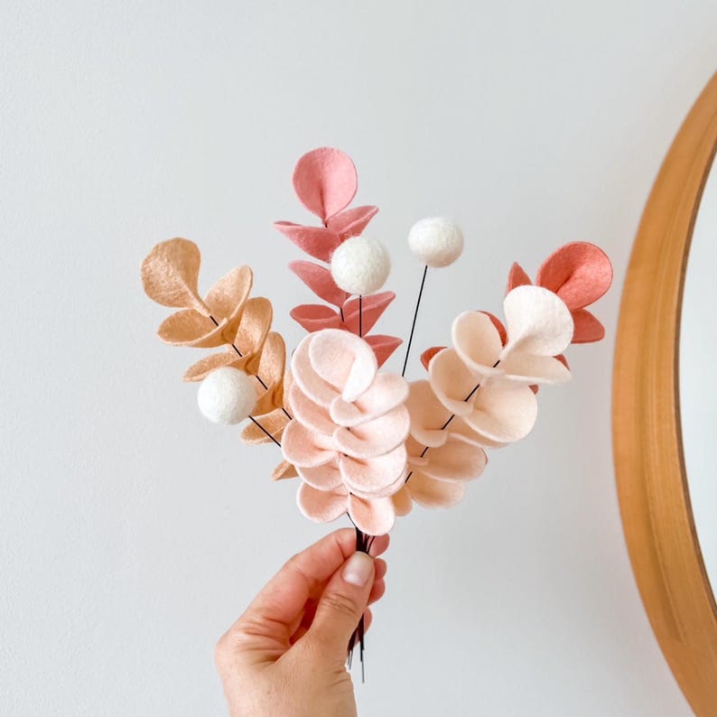 Pode incluir: Um ramo decorativo de hastes de feltro com folhas arredondadas em tons de p&ecirc;ssego, creme e rosa seco, intercaladas com pequenas esferas de feltro branco presas a hastes de arame fino e escuro, dispostas num grupo para fins decorativos.