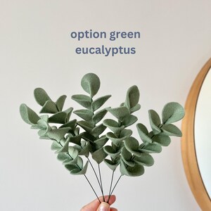 May include: A decorative eucalyptus branch made of sage green felt with rounded leaves attached to thin black wire stems. The text option green eucalyptus is printed in blue above the plant. The felt leaves have a soft matte texture.