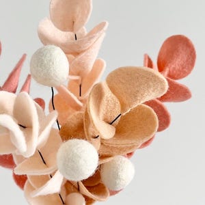 Pode incluir: Um arranjo floral decorativo feito de feltro, com folhas arredondadas em tons de creme, p&ecirc;ssego e terracota. Os caules s&atilde;o intercalados com pequenas esferas de feltro branco s&oacute;lido, criando uma composi&ccedil;&atilde;o bot&acirc;nica texturizada e em camadas.