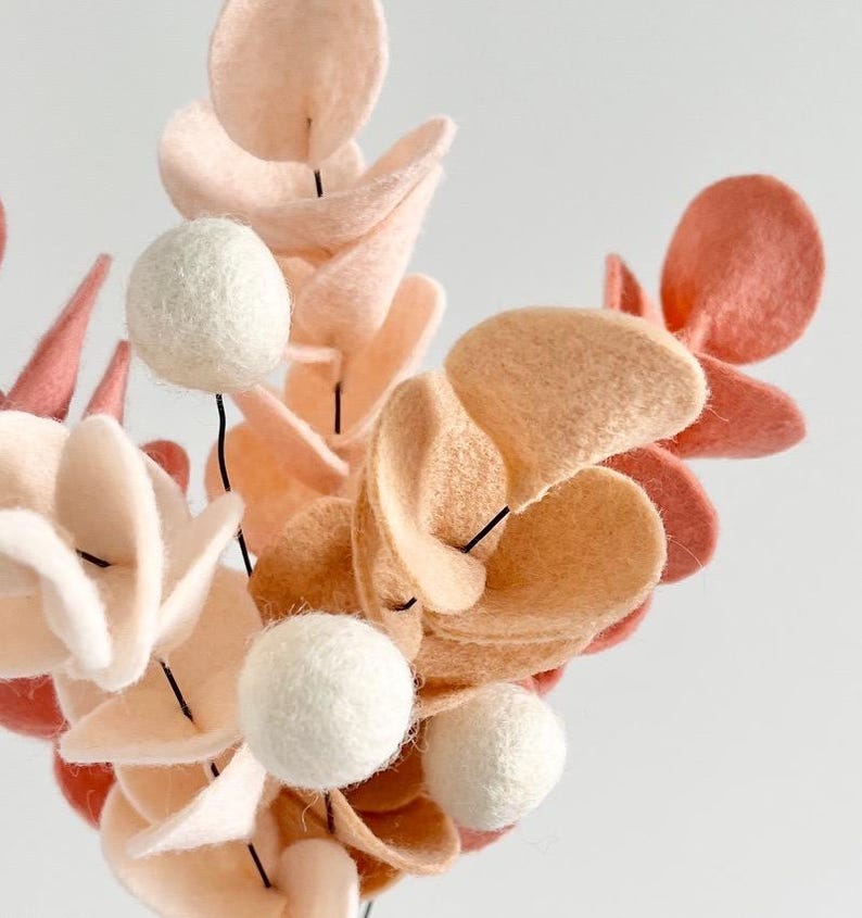 Pode incluir: Um arranjo floral decorativo feito de feltro, com folhas arredondadas em tons de creme, p&ecirc;ssego e terracota. Os caules s&atilde;o intercalados com pequenas esferas de feltro branco s&oacute;lido, criando uma composi&ccedil;&atilde;o bot&acirc;nica texturizada e em camadas.