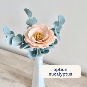 Puede incluir: Un arreglo floral de fieltro con una peon&iacute;a color melocot&oacute;n con un centro amarillo, rodeada de hojas de eucalipto azul claro. El arreglo est&aacute; en un jarr&oacute;n blanco. El texto en la imagen dice "option eucalyptus".