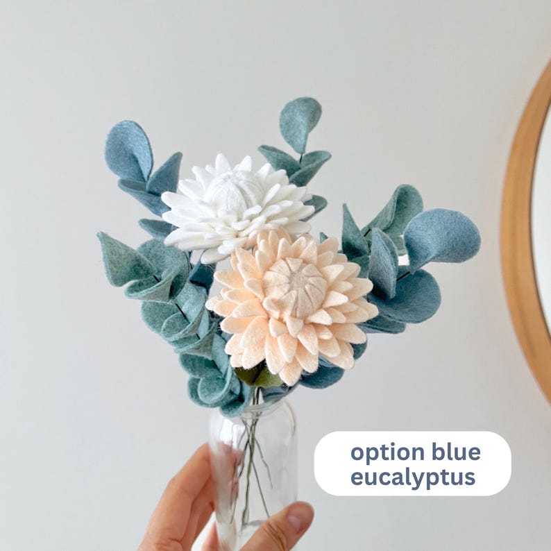 Op de afbeelding: Een boeket van viltbloemen in een doorzichtige glazen vaas, met &eacute;&eacute;n witte en &eacute;&eacute;n licht perzikkleurige bloem omringd door blauwgroene vilten eucalyptusbladeren. De tekst op de afbeelding luidt option blue eucalyptus.