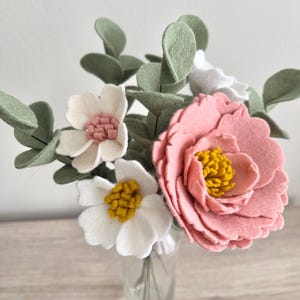 May include: A bouquet of felt flowers featuring a large pink peony with a yellow center, two white flowers with yellow and pink centers, and several sage green felt eucalyptus leaves arranged in a clear glass vase.