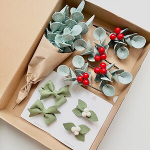May include: A cardboard box containing a bouquet of felt eucalyptus leaves wrapped in brown paper, three sprigs of felt eucalyptus with red berries, two sage green felt bows, and two hair clips featuring felt leaves and white pom-poms.