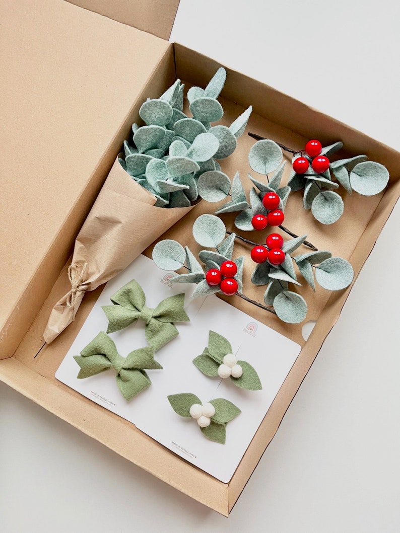 May include: A cardboard box containing a bouquet of felt eucalyptus leaves wrapped in brown paper, three sprigs of felt eucalyptus with red berries, two sage green felt bows, and two hair clips featuring felt leaves and white pom-poms.