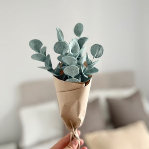 May include: A small bouquet of felt eucalyptus stems with rounded sage green leaves, wrapped in brown kraft paper and tied with a simple string. The stems are arranged in a vertical cluster, showcasing the soft texture of the felt material.