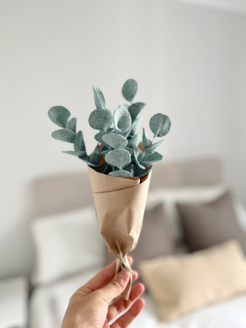 May include: A small bouquet of felt eucalyptus stems with rounded sage green leaves, wrapped in brown kraft paper and tied with a simple string. The stems are arranged in a vertical cluster, showcasing the soft texture of the felt material.