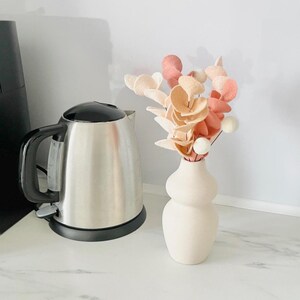 Pode incluir: Um vaso de cer&acirc;mica cor de creme mate com formato arredondado de ampulheta contendo um buqu&ecirc; de flores de feltro em tons de p&ecirc;ssego, coral e branco, posicionado ao lado de uma chaleira el&eacute;trica de a&ccedil;o inoxid&aacute;vel com al&ccedil;a preta.