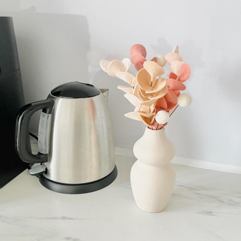 Pode incluir: Um vaso de cer&acirc;mica cor de creme mate com formato arredondado de ampulheta contendo um buqu&ecirc; de flores de feltro em tons de p&ecirc;ssego, coral e branco, posicionado ao lado de uma chaleira el&eacute;trica de a&ccedil;o inoxid&aacute;vel com al&ccedil;a preta.