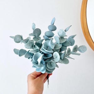 May include: A decorative bouquet of artificial eucalyptus stems featuring rounded, felt-like leaves in a muted, dusty blue-green color. The stems are gathered together at the base, showcasing a textured, matte finish on each individual leaf.