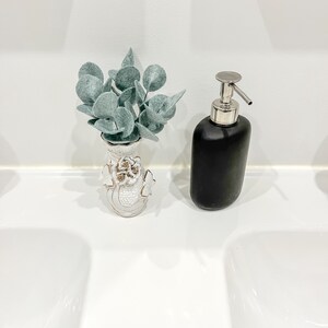 May include: A small white ceramic vase with gold-trimmed floral relief details containing artificial sage green eucalyptus leaves, positioned next to a matte black soap dispenser with a silver-toned metal pump mechanism.