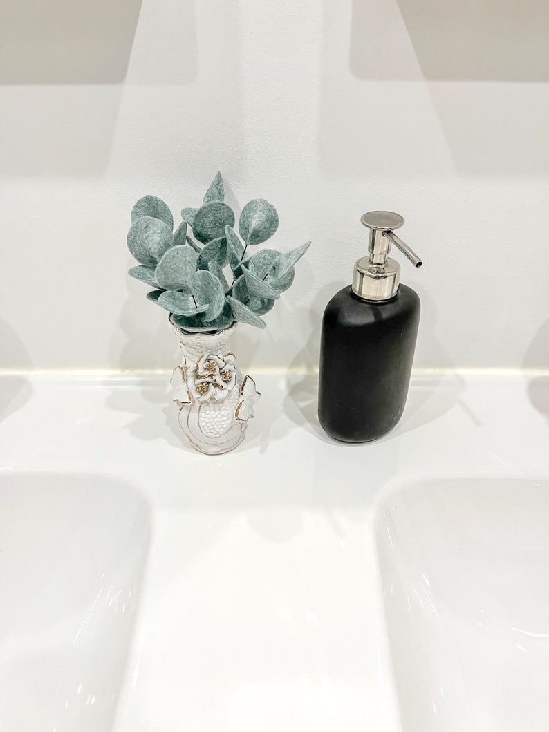 May include: A small white ceramic vase with gold-trimmed floral relief details containing artificial sage green eucalyptus leaves, positioned next to a matte black soap dispenser with a silver-toned metal pump mechanism.