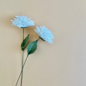 May include: Two white felt flowers with layered petals attached to thin green stems. Each stem features a single pointed green leaf. The flowers are positioned at an angle, showcasing the white texture of the felt and the green stems.