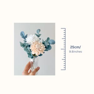 May include: A bouquet of felt flowers in a clear glass vase, featuring one white flower, one pale peach flower, and several blue-green eucalyptus-style leaves. The arrangement measures 9.8 inches in height as indicated by the adjacent scale.