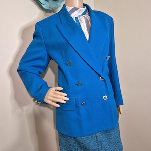Puede incluir: Un blazer azul vibrante de doble botonadura con botones dorados y una falda azul a juego. El blazer presenta un diseño de solapa clásico y un ajuste a medida. El maniquí también lleva una bufanda a rayas y un sombrero negro.