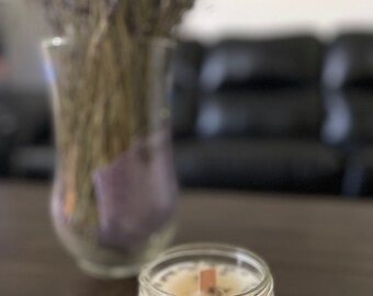 Puede incluir: Una vela blanca con una mecha de madera en un tarro de vidrio transparente. La vela tiene botones de lavanda secos incrustados en la cera.