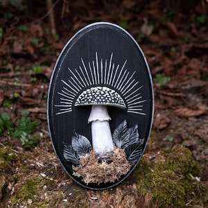 May include: Black oval wooden wall art featuring a detailed mushroom design. The mushroom has a white stem and a speckled cap, with white rays emanating from behind it. The artwork includes carved leaves and moss.