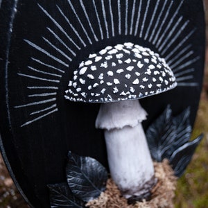 May include: A black and white mushroom sculpture with a detailed cap covered in white spots. The mushroom is set against a black background with white radiating lines, and black leaves. The sculpture is placed on a bed of brown moss.