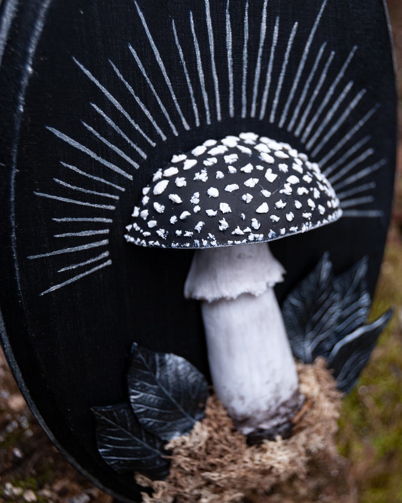May include: A black and white mushroom sculpture with a detailed cap covered in white spots. The mushroom is set against a black background with white radiating lines, and black leaves. The sculpture is placed on a bed of brown moss.