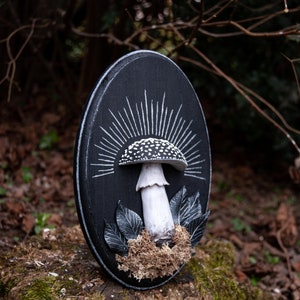 May include: A black, oval-shaped wooden plaque featuring a detailed mushroom sculpture. The mushroom has a white stem and a cap with black spots. White rays emanate from behind the mushroom, and there are decorative leaves and moss.