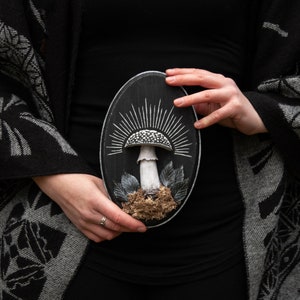 May include: A black oval wooden plaque featuring a detailed mushroom design. The mushroom has a white and black spotted cap, a white stem, and is surrounded by leaves and moss. White rays emanate from behind the mushroom.
