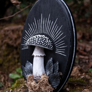 May include: A black oval wooden plaque featuring a white and black mushroom design. The mushroom has a white stem and a cap with white spots. White rays emanate from behind the mushroom. Gray leaves and brown moss are at the base.