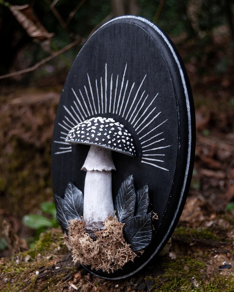 May include: A black oval wooden plaque featuring a white and black mushroom design. The mushroom has a white stem and a cap with white spots. White rays emanate from behind the mushroom. Gray leaves and brown moss are at the base.