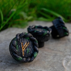 Op de afbeelding: Close-up van drie donkere, getextureerde decoratieve knoppen. De knoppen hebben een zwart-groen kleurenschema met een goudkleurig rune-symbool. De knoppen zijn gerangschikt op een grijs stenen oppervlak met groen gras op de achtergrond.