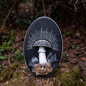 May include: Oval black wooden plaque featuring a white and black mushroom design with radiating lines. The mushroom has a white stem and a dotted cap. Gray leaves and brown moss surround the mushroom. The plaque is displayed outdoors.