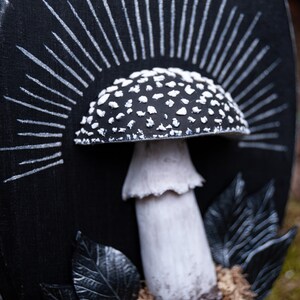 May include: A black and white mushroom sculpture with a detailed cap covered in white spots. The mushroom is set against a black background with white radiating lines, and is surrounded by black leaves and moss.