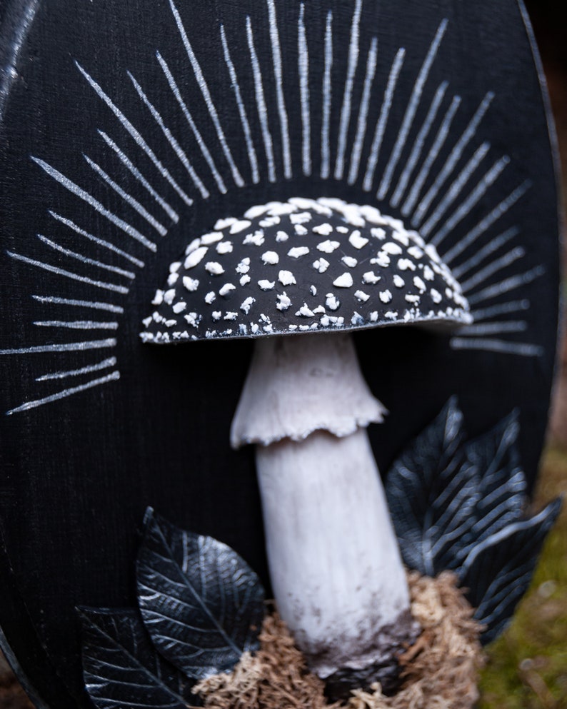 May include: A black and white mushroom sculpture with a detailed cap covered in white spots. The mushroom is set against a black background with white radiating lines, and is surrounded by black leaves and moss.