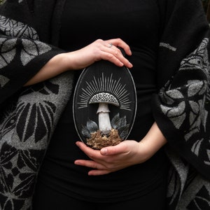 May include: A black oval wooden plaque featuring a detailed mushroom sculpture. The mushroom has a white stem and a speckled cap, surrounded by leaves and moss. The background is a sunburst design.