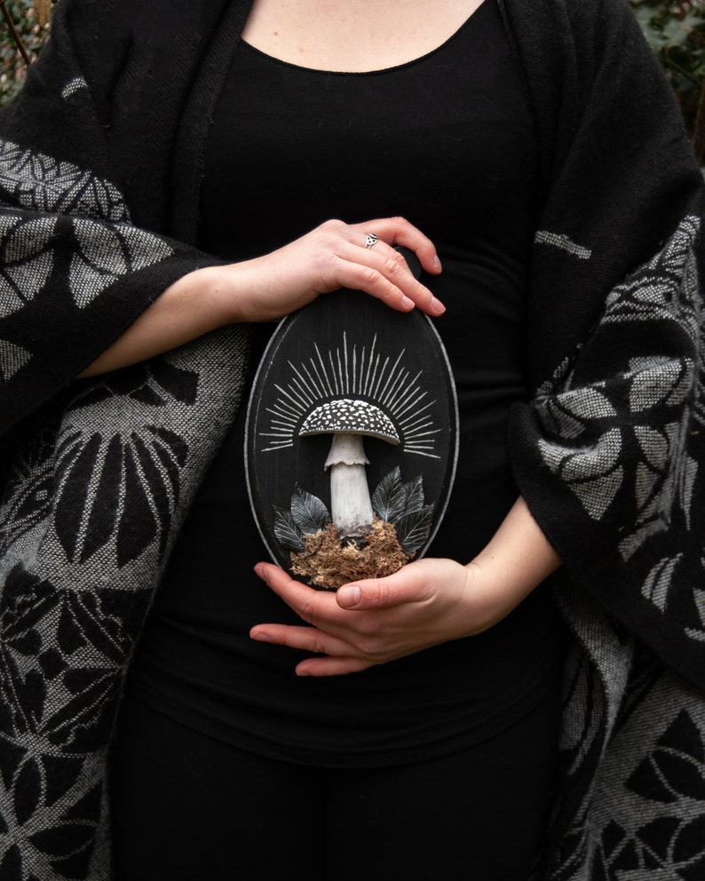 May include: A black oval wooden plaque featuring a detailed mushroom sculpture. The mushroom has a white stem and a speckled cap, surrounded by leaves and moss. The background is a sunburst design.