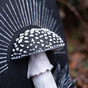 May include: A black and white mushroom sculpture with a sunburst design. The mushroom cap is black with white spots, and the stem is white. The background is a dark, textured surface.