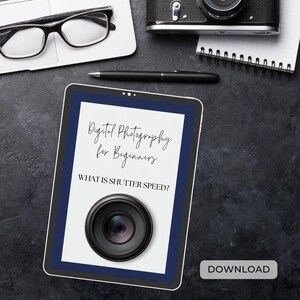 May include: A digital tablet displaying a white page with the text "Digital Photography for Beginners" and "WHAT IS SHUTTER SPEED?" with a black and silver camera lens illustration. The tablet has a blue border and the word "DOWNLOAD" is printed in white on a blue button at the bottom of the screen.