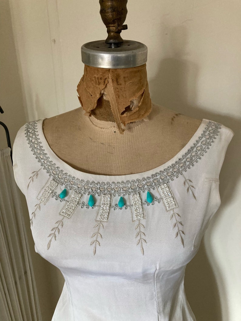 May include: A white sleeveless top featuring a wide, rounded neckline adorned with intricate silver metallic embroidery, delicate leaf-patterned stitching, and four bright turquoise teardrop-shaped embellishments centered along the collar.