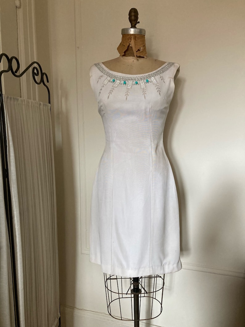 May include: A sleeveless white dress featuring a round neckline adorned with silver embroidery, clear rectangular beads, and small turquoise-colored stones. The dress has a fitted bodice with vertical seams extending down to the hemline.