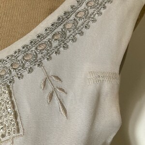 May include: A cream-colored sleeveless garment featuring a textured, ribbed fabric. The neckline is adorned with intricate silver metallic embroidery and small circular accents. Additional embroidered leaf motifs and a rectangular beaded panel are visible.