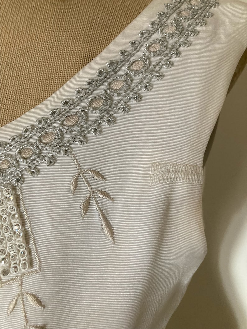 May include: A cream-colored sleeveless garment featuring a textured, ribbed fabric. The neckline is adorned with intricate silver metallic embroidery and small circular accents. Additional embroidered leaf motifs and a rectangular beaded panel are visible.