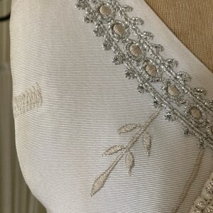 May include: A white garment featuring a textured ribbed fabric surface. The piece is adorned with an intricate silver metallic embroidered trim along the neckline, complemented by a delicate embroidered leaf motif and a row of small white beads.