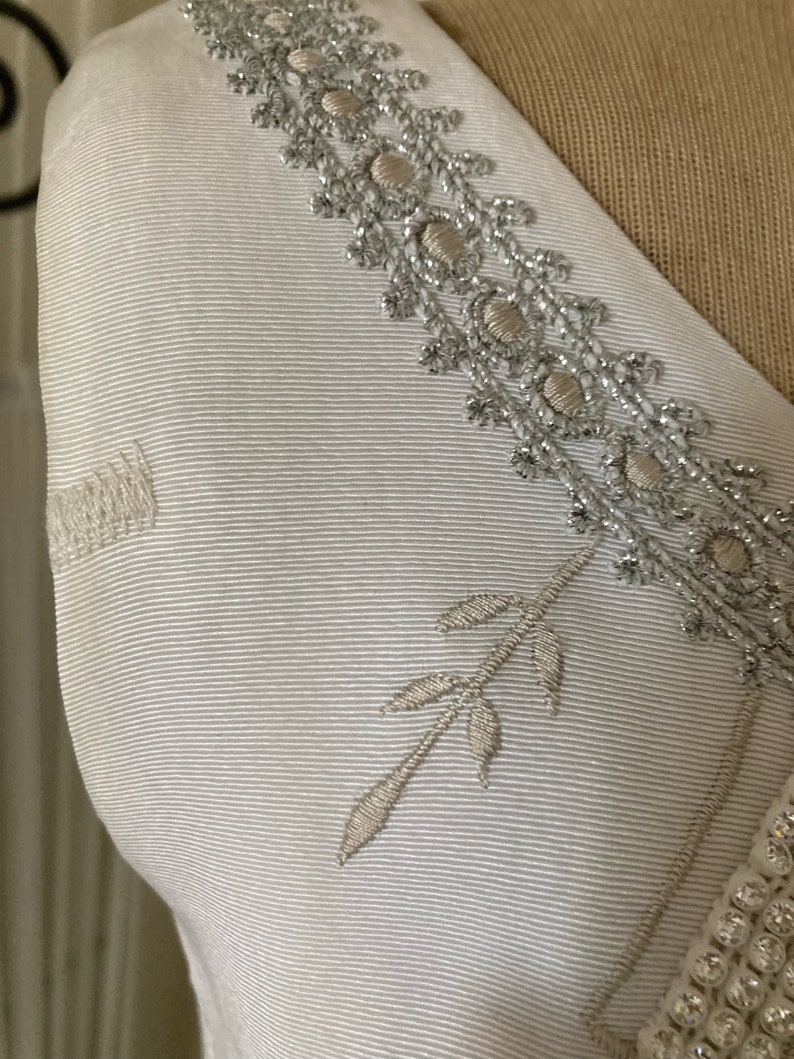 May include: A white garment featuring a textured ribbed fabric surface. The piece is adorned with an intricate silver metallic embroidered trim along the neckline, complemented by a delicate embroidered leaf motif and a row of small white beads.